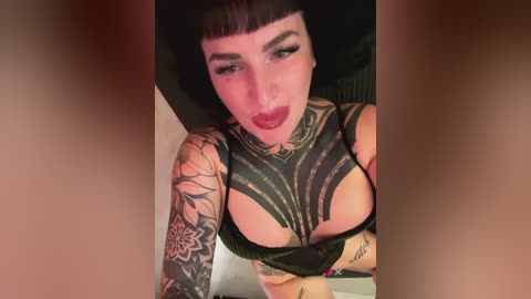 Media: Video of a tattooed woman with dark hair and bold makeup, wearing black lingerie, revealing her large breasts. She stands indoors with a blurred background.