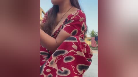 Media: Video of a woman with medium skin tone and long dark hair, wearing a red dress with a pattern of white and black leaf shapes. She is outdoors, next to a building with yellow walls and a blue sky in the background.