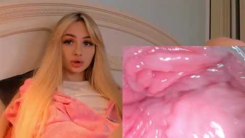 Media: Video of a young woman with long blonde hair, fair skin, wearing a white shirt under a pink blanket, with a close-up of her vaginal area showing pink, moist tissue.