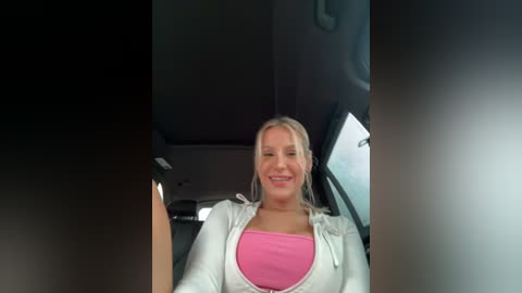 Media: Video of a smiling, light-skinned woman with blonde hair, wearing a pink top and white zip-up jacket, seated in a car with a dark interior.