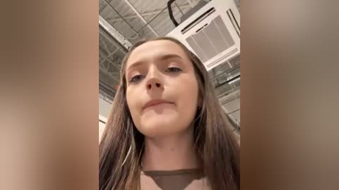 Media: Video of a young, fair-skinned woman with long, straight brown hair, wearing a beige top, looking down from a high angle, taken indoors with a metal ceiling and industrial elements visible in the background.