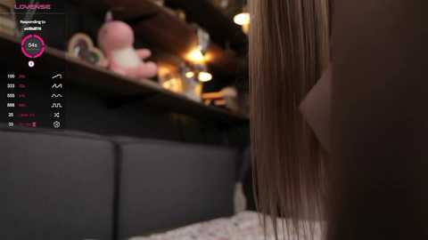 Media: A blurry video of a nude woman with long, straight blonde hair, standing near a dark sofa and a shelf with plush toys, in a dimly lit room.