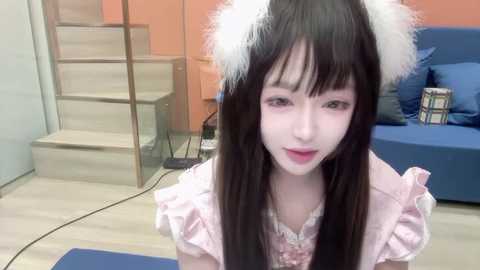 Media: A video of an Asian woman with pale skin, long black hair, and pink bunny ears, wearing a pink ruffled dress, sitting on a blue couch. Background includes stairs and orange wall.