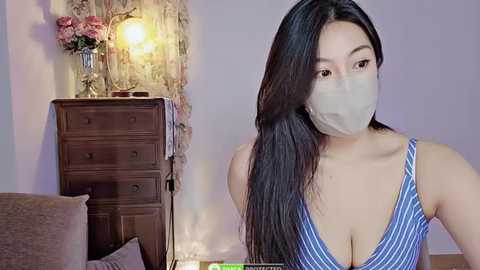 Media: Video of an East Asian woman with long black hair, wearing a blue and white striped tank top, face mask, and sitting indoors. Background includes a wooden dresser, vase, and floral patterned curtains.