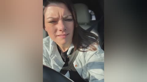 Media: Video of a woman wearing a white shirt and a black tie, driving with a grim expression. She has long brown hair and is seated in a car, with the car seat visible in the background.