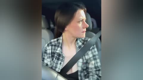 Media: Video of a woman with shoulder-length brown hair, wearing a black-and-white plaid shirt, buckled into a car seat. She has a neutral expression, with a blurred background showing car interior.