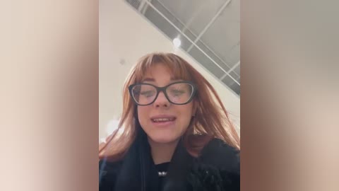 Media: Video of a young woman with fair skin and red hair, wearing black-framed glasses and a black jacket, standing in a modern, brightly lit room with a high ceiling.