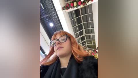 Media: Video of a young woman with red hair and glasses, wearing a black fur coat, standing indoors with floral decorations and a window in the background.