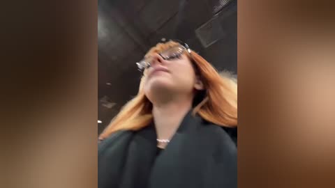 Media: Video of a woman with long, straight, reddish-brown hair, wearing glasses, a black coat, and a white necklace, standing indoors with a blurred background.