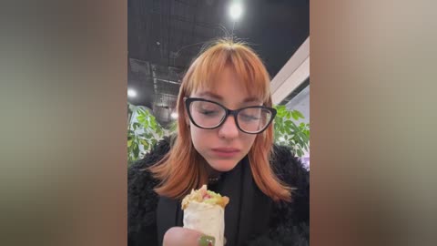 Media: Video of a young woman with straight, shoulder-length auburn hair, wearing black-framed glasses and a black fur coat, eating a taco with lettuce and cheese. Background includes green plants and a modern, dimly lit interior.