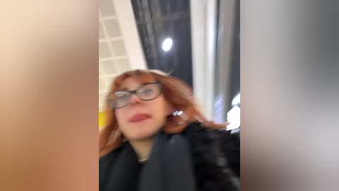 Media: Video of a fair-skinned woman with red hair, wearing glasses and a black leather jacket, standing in an indoor setting with blurred background, likely a subway station.