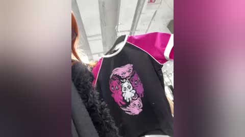 Media: Video of a black and pink hoodie with a graphic of a smiling skull inside a black circle, hanging on a wooden hanger. Background shows a blurred interior with white walls and metal structures.