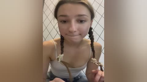 Media: A video of a young woman with braided hair, wearing a white tank top, kneeling in front of a white diamond-patterned wall, with her hands tied in front.