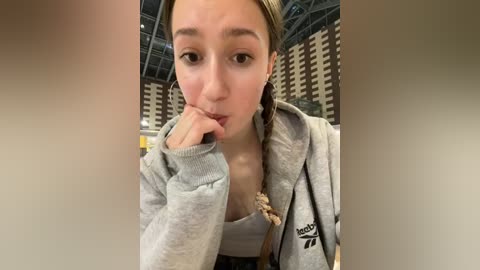 Media: A video of a young woman with light skin and blonde braided hair, wearing a grey sweatshirt, taking a selfie in a sports stadium with rows of seats in the background.