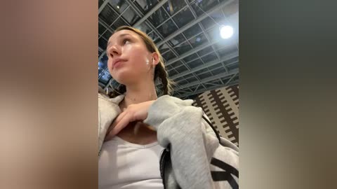 Media: Video of a young woman with light skin and brown hair, wearing a white top and gray hoodie, looking upward. Background features a ceiling with metal beams and a patterned wall.