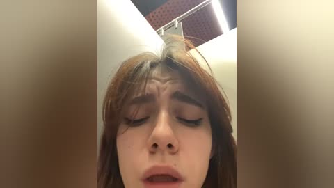 Media: A video of a young woman with fair skin and shoulder-length, auburn hair, eyes closed, and mouth slightly open, taken from a low angle. The background is an indoor space with beige walls and a perforated ceiling.