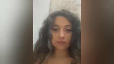 Media: Video of a young woman with long, curly black hair and light skin, looking directly at the camera. She has a neutral expression. Background features a light-colored wall and a textured beige pillow.