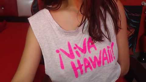 Media: Video of a woman with medium-length brown hair wearing a light gray sleeveless tank top with \"Neva Hawa\" in bold pink letters. The background features a red carpet and a blurred, colorful fabric.