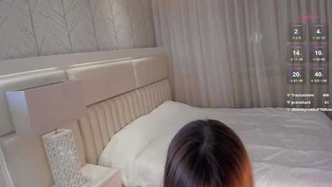 Media: Video of a modern bedroom with a beige tufted headboard, white bedding, and soft, patterned wallpaper. A person with long, dark hair lies on the bed, partially visible from the back. A digital screen in the corner displays live stream details.