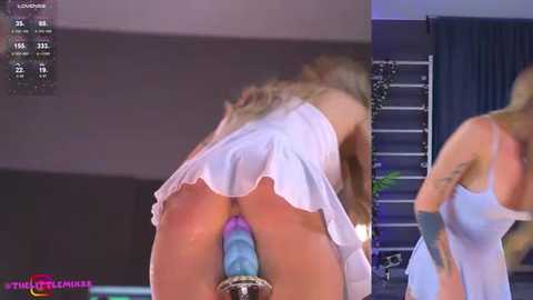 Media: Video of a blonde woman bent over, wearing a white, ruffled dress, exposing her buttocks with a large, colorful dildo inserted. Background includes a dark room with a silver ladder and a blue curtain.