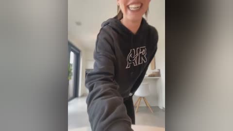 Media: Video of a smiling woman with light skin and dark hair, wearing a black hoodie with white text, standing in a modern, brightly lit kitchen with white cabinets and a glass door.