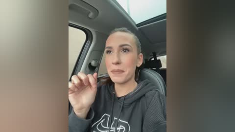 Media: Video of a young woman with light skin, brown hair tied back, wearing a black hoodie, sitting in a car with gray interior, looking serious.