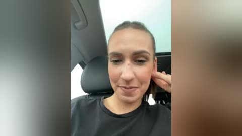 Media: Video of a woman with light skin and dark hair, wearing a black cape, sitting in a car, adjusting her earpiece.