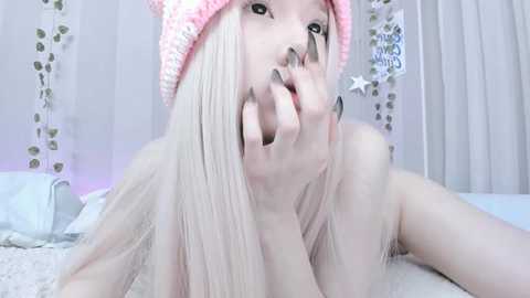 Media: Video of a pale-skinned woman with long blonde hair, wearing a pink knit hat, topless, biting her fingers, in a pastel-themed bedroom with floral wallpaper.