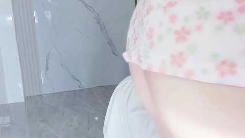 Media: Video of a woman's upper torso, wearing a white floral-patterned crop top and white pants, standing in a bathroom with marble walls and grey tiled floor.