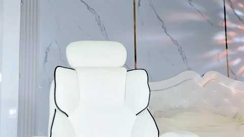 Media: Video of a plush, white ergonomic office chair with a high backrest, set in a minimalist room with a reflective, marble-patterned wall and a white, tufted headboard.