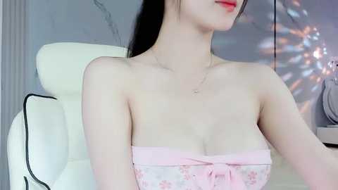 Media: A video of a young Asian woman with pale skin, black hair, wearing a strapless pink floral dress, seated on a white leather chair. Background includes a gray wall with a decorative, reflective object.