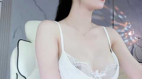 Media: Video of a slender, light-skinned woman with long black hair, wearing a white lace-trimmed bra, sitting in a white leather chair against a marble wall.