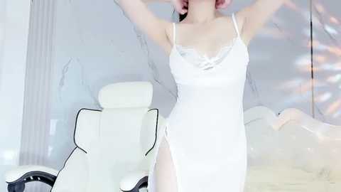 Media: A video of a slim, fair-skinned woman in a white lace nightgown, stretching with arms raised, in a modern, minimalist room with white walls, a white leather chair, and a bed with white bedding.