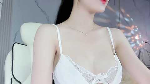 Media: Video of a fair-skinned woman with long black hair, wearing a white lace-trimmed spaghetti strap top, sitting in a white chair, against a marble wall.