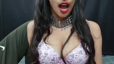 Media: Video of a woman with long black hair, wearing a lace bra with a pink floral pattern, an off-the-shoulder olive-green top, and a silver chain necklace, mouth open in an expression of surprise or excitement, against a dark background.