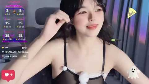 Media: A young Asian woman with long black hair, fair skin, and a slim physique, wearing a sheer black lingerie top with fluffy white pom-poms, sits indoors, smiling.