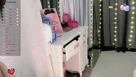 Media: Video of a chic, pastel-toned dressing room with white vanity, pink shelves, and a pink hamper; soft, glowing fairy lights adorn the background.