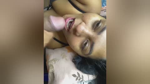 Media: A close-up video of a woman with medium brown skin and dark hair, lying on a bed, performing oral sex on a penis. She has a relaxed expression. The background includes floral-patterned sheets.