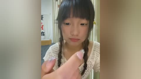 Media: Video of an Asian woman with long black hair in braids, wearing a white crochet top, holding a small, uncircumcised penis in her hand. Background shows a beige wall and a wooden table with red and black decorations.