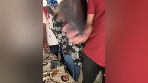 Media: Video of a woman in a black floral dress with blue jeans, being held by a person in a red shirt and black pants, in a cluttered room with a colorful patterned bedspread.