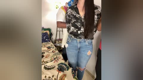 Media: Video of a young woman with long dark hair, wearing a black floral blouse and light blue distressed jeans, standing in a cluttered bedroom with a bed covered in a colorful, patterned duvet.