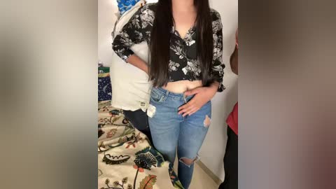 Media: Video of a young woman with long, straight black hair, wearing a black floral top and ripped blue jeans, standing in a bedroom with a floral-patterned bedspread.