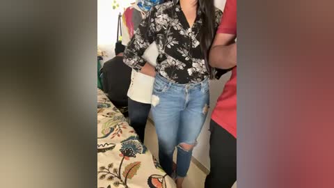 Media: Video of a woman with medium skin tone wearing a floral blouse and ripped jeans, standing in a cluttered bedroom with colorful bedspread.