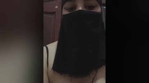 Media: Video of a woman wearing a black face mask, revealing only her eyes, with a beige top and a dark background.