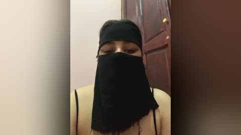 Media: Video of a woman with medium skin tone wearing a black face mask, black headband, and a beige top, standing indoors near a brown door with a yellow light on.