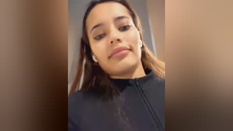 Media: A close-up video of a young woman with medium brown skin and straight, shoulder-length dark hair, wearing a black zip-up jacket. Her expression is neutral, and she is listening to white earbuds.