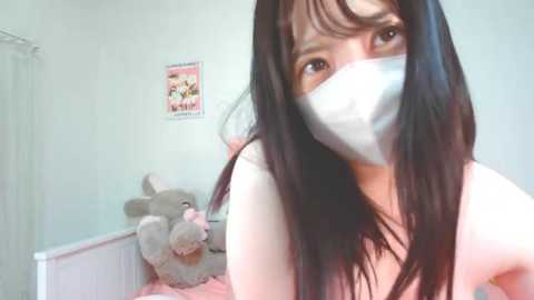 Media: Video of a young Asian woman with long black hair, wearing a pink mask and a light pink top, in a pastel room with a stuffed rabbit and a framed poster.