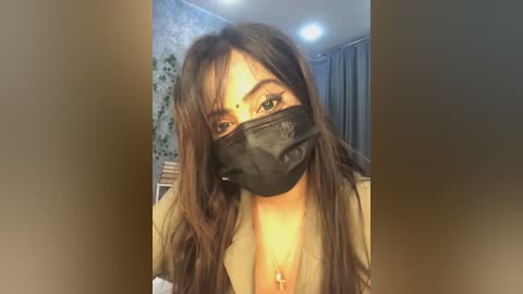 Media: Video of a young woman with long, dark hair, wearing a black face mask, tan blazer, and a gold cross necklace. She has light skin and a slender build. Background shows a floral wallpaper and a bed with a gray curtain.