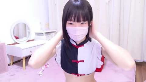 Media: Video of a young East Asian woman with straight black hair, wearing a white and black maid cosplay outfit with red accents, face mask, and gloves, kneeling on a pink carpeted floor in a brightly lit room with a vanity table in the background.