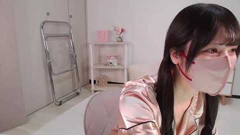 Media: Video of an Asian woman with long black hair, wearing a white surgical mask and a pink satin robe, kneeling on a white chair in a minimalist, sterile room with a metal folding chair and a white shelf in the background.